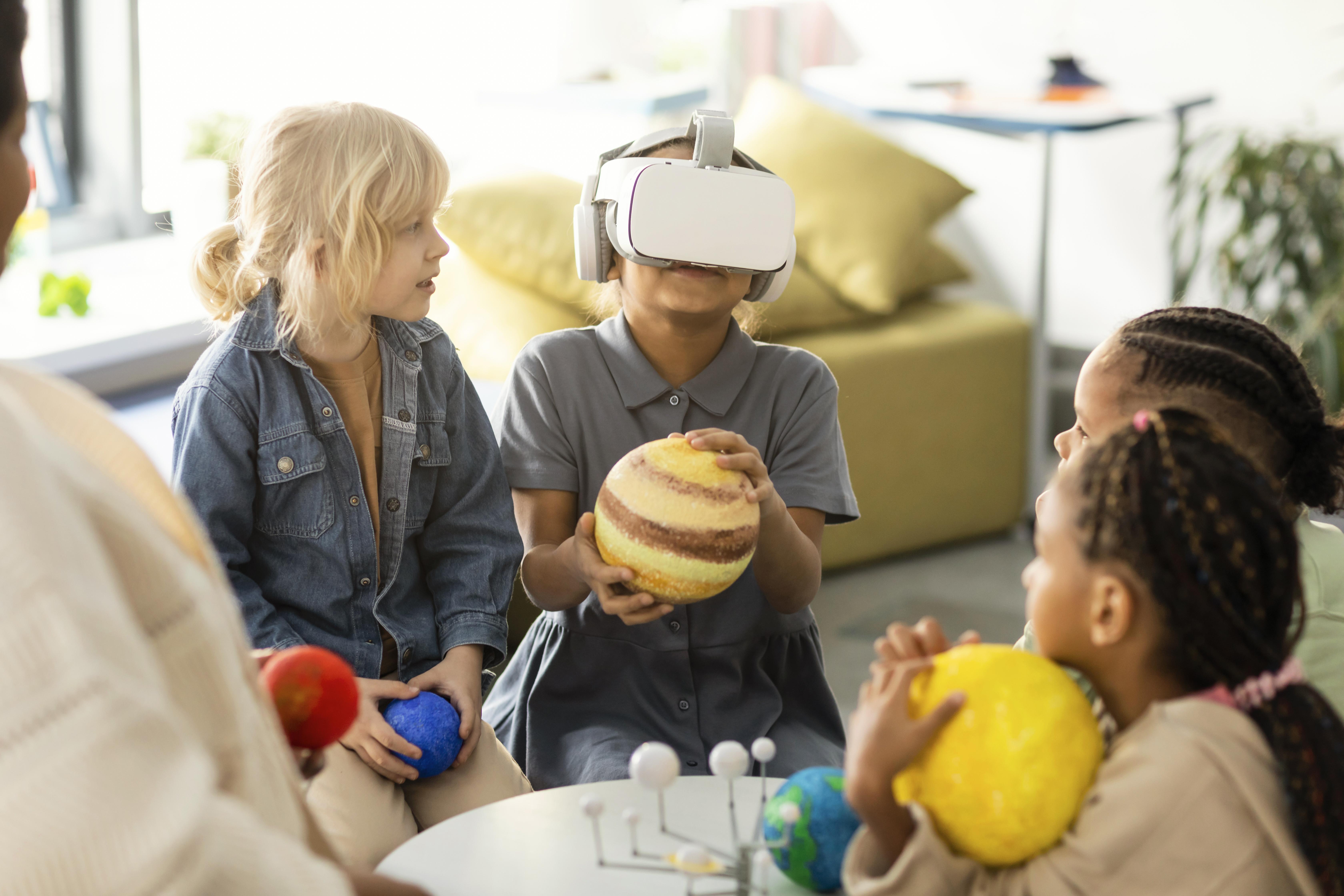 Várias crianças estão sentadas em círculo, interagindo com modelos de planetas e um óculos de realidade virtual.
